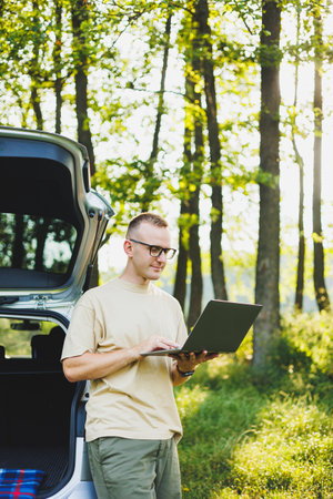 Cheerful traveler, young man sitting in open trunk of car, working with laptop, outdoors, concept of...