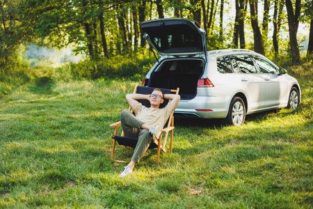 A cheerful man sits on a chair in nature and works online on a laptop. A man travels and works remotely on a laptop computer. Office work in natureの写真素材