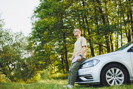 A successful man in glasses works on a laptop while leaning on the hood of his car. Remote work in...