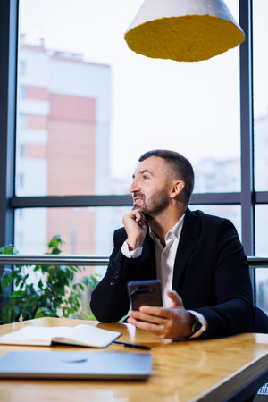Confident male businessman in a business suit sits at a table with a phone and works remotely