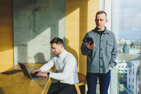 Two confident young people are sitting together at a table and one of them is pointing at a laptop....