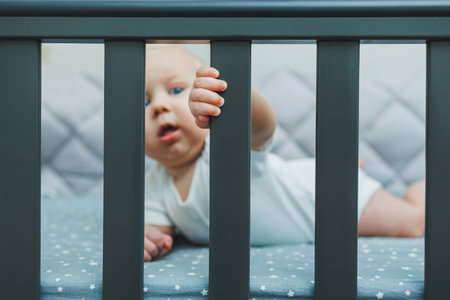 The baby looks through the sides of the crib into the room. A small newborn boy lies in a crib for children. A place for children to sleepの写真素材