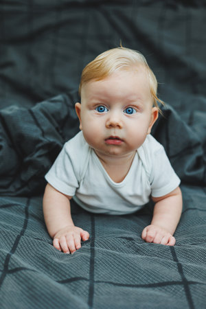 A cheerful baby boy with beautiful big eyes is lying on the bed at home in a white bodysuit on gray...