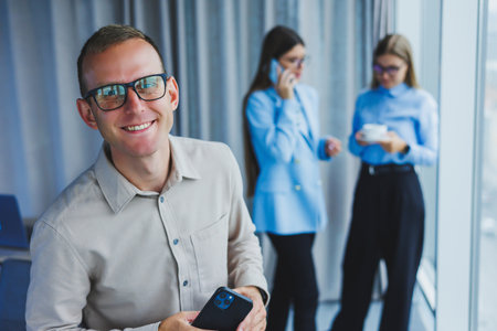 Smiling young male manager in glasses talking on the phone while standing in the office with colleagues in the background. A working day in a modern office with large windowsの写真素材