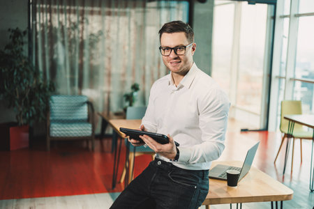 A young successful man in glasses and a white shirt is working on a tablet on a project in a modern...