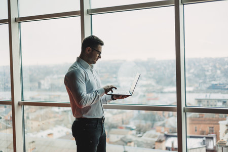 A young handsome man with a laptop stands on the background of a large panoramic window in the sky....