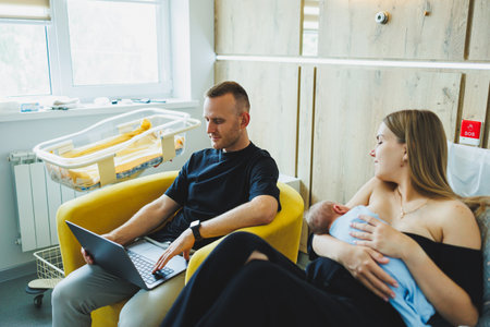 Remote online work. A happy father is working on a laptop while his wife is taking care of the child. Working from home, parenting and remote work. Breastfeeding a newborn babyの写真素材