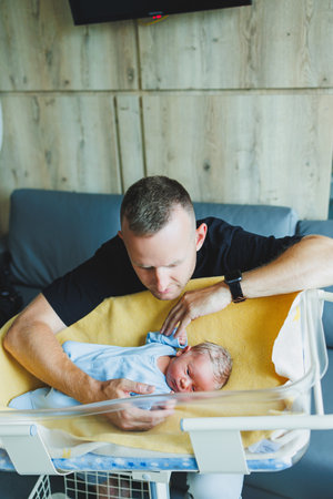 A cute young father is sitting over the crib of his newborn son. A father hugs a newborn baby.