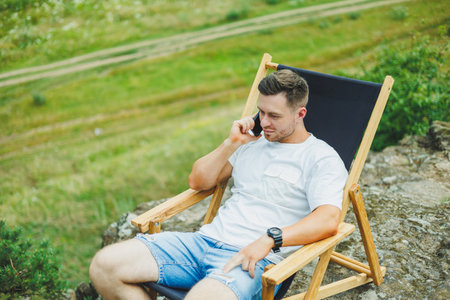 A bearded man sits on a folding wooden chair in nature and talks on the phone. The concept of recreation in nature alone.の写真素材