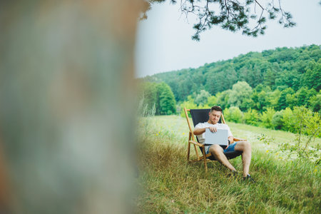 A happy man is relaxing in nature and working remotely on a portable laptop. Remote work while on...