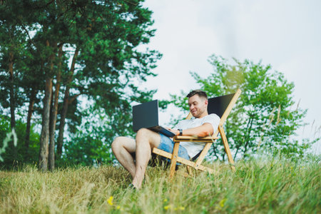 A man on a folding wooden chair sits in nature and works remotely online on a laptop. The concept of recreation in nature alone.の写真素材