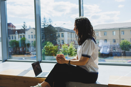 Cheerful woman sitting with laptop, smartphone, coffee cup, looking at laptop and taking notes while...
