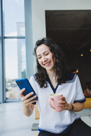 Freelance work. A young smiling curly-haired woman sits near the large windows in a cafe and works...