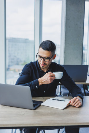 A handsome manager man of Caucasian appearance in a black shirt is sitting at a table and drinking...