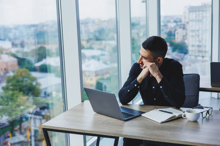 A smiling businessman in a black shirt is sitting at a desk using a laptop. A person works in the office on a computer, analyzes data. Internet marketing, adult education, e-learning.の写真素材
