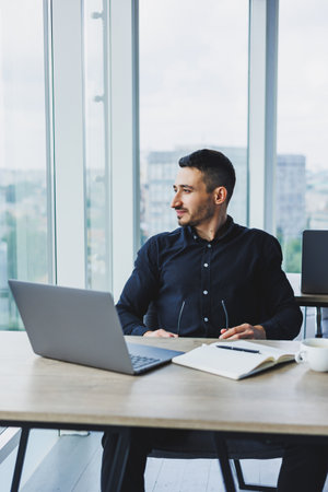 A smiling businessman in a black shirt is sitting at a desk using a laptop. A person works in the...