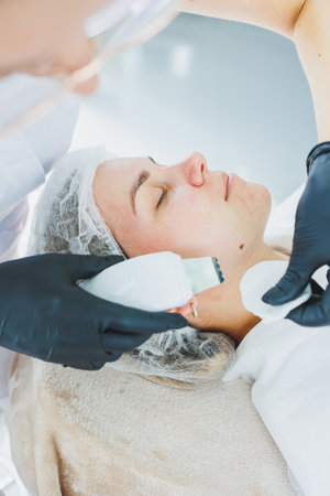 A dermatologist or cosmetologist in a clinic works with female patients and makes facial skin procedures on a young woman's face. Ultrasonic cleaning of the face in the cosmetologist's officeの写真素材