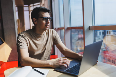 Young handsome smiling smart freelancer in glasses works at a laptop while spending time in a cafe....