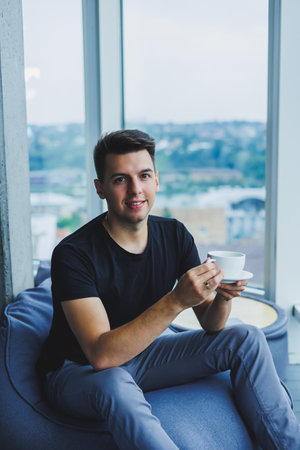 A young man sits on a bean bag chair near a large window in the office and drinks coffee during his...