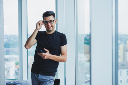 A successful young businessman in glasses is writing a message on the phone near the window in a...