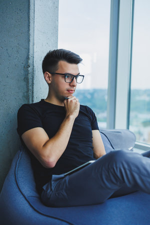 A young male student of Caucasian appearance sits in a bag chair and takes notes in a notebook. The...
