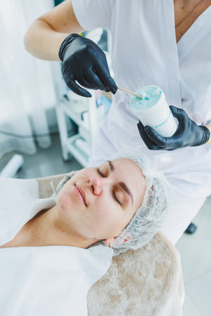 A beautician applies a mask to a woman's face. Facial skin care. A dermatologist or cosmetologist in a clinic works with female patients and makes facial skin procedures on a young woman's face.の写真素材
