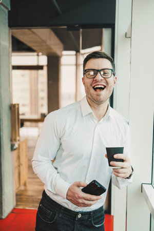 A male manager in a white shirt and glasses stands in the office by the window and drinks coffee while watching a video on the phone. Business telephone conversation of a business man.の写真素材