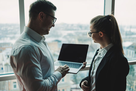 A man and a woman are standing in the office against the background of a large panoramic window. Modern spacious workspace with employees. Business partnersの写真素材