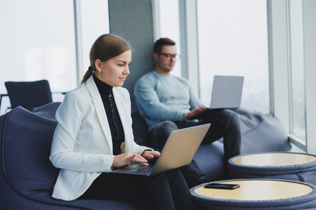 Young colleagues sit on soft poufs and work remotely on laptops. Modern workspace with large windows.の写真素材