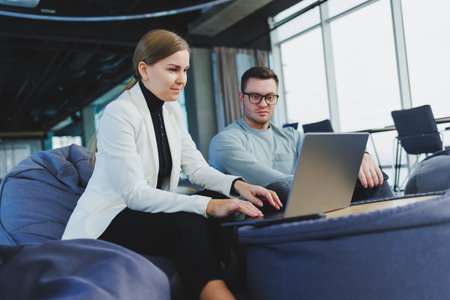 Young colleagues sit on soft poufs and work remotely on laptops. Modern workspace with large windows.の写真素材