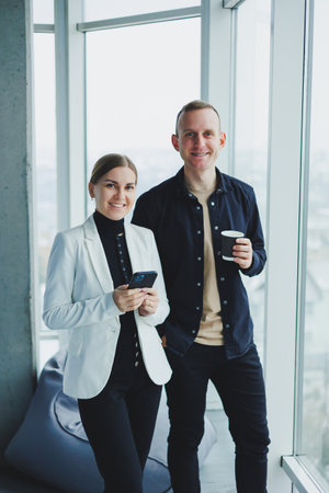 Smiling man and woman standing near window with gadgets in modern transparent office. Conversation of business colleagues in an informal setting.の写真素材
