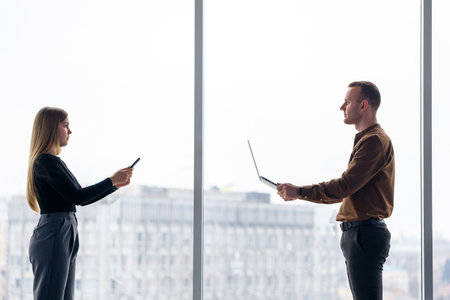 A successful female office worker with a colleague with a netbook stands in a skyscraper against the backdrop of a window overlooking the city. City architects looking at laptop happy with projectの写真素材