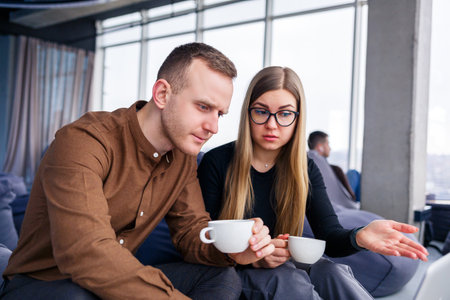 A successful young woman manager with her boss sits with a laptop on an upholstered armchair by the panoramic window and drinks coffee. Business woman and man working on a new projectの写真素材