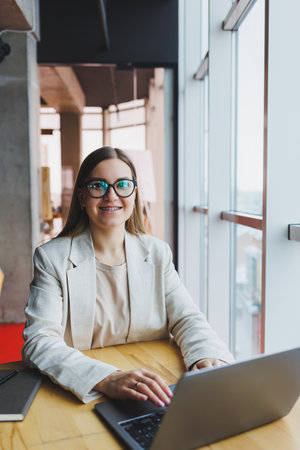 Successful happy young woman in jacket smiling and working on laptop in modern office with large windows. Remote workの写真素材
