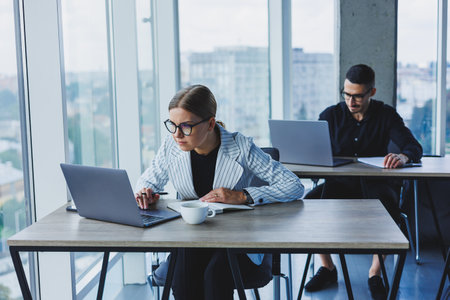 Working atmosphere in an office with large windows. A female manager is sitting at a desk using a laptop in a modern office against the background of a colleague.の写真素材