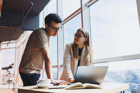 Two colleagues, a man and a woman manager, stand near a table with a laptop in a modern office and discuss a new project. Two business partners in glasses are talking about a new business.の写真素材