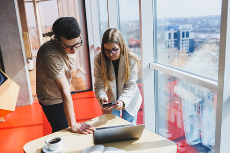 Two colleagues, a man and a woman manager, stand near a table with a laptop in a modern office and...