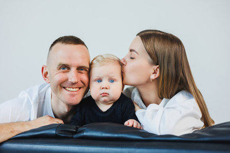 A young married couple with a small child is photographed sitting on a sofa on a white background. Caring parents look at their baby while holding it in their arms.の写真素材