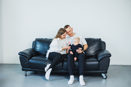 A young married couple with a small child is photographed sitting on a sofa on a white background. Caring parents look at their baby while holding it in their arms.の写真素材