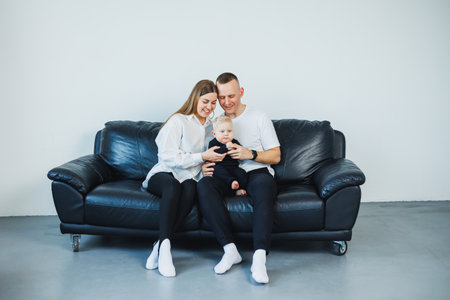 A young married couple with a small child is photographed sitting on a sofa on a white background. Caring parents look at their baby while holding it in their arms.の写真素材