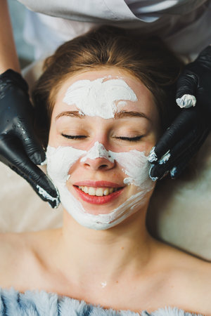 The hands of a beautician apply a mask to a woman's face in a spa salon. Aesthetic procedure. Facial skin care.の写真素材
