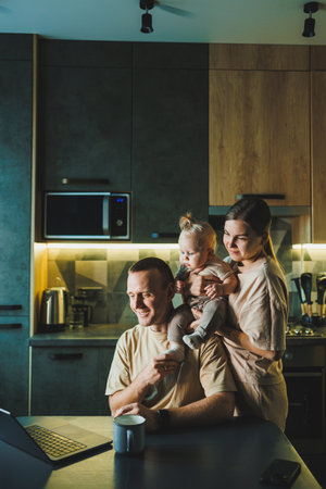 A young father works on a laptop while his wife takes care of the child. Work from home. A young couple in the kitchen with a childの写真素材