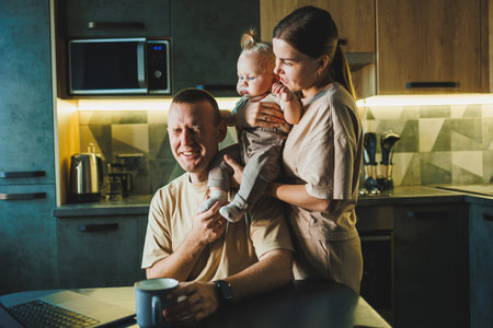 A young father works on a laptop while his wife takes care of the child. Work from home. A young couple in the kitchen with a childの写真素材