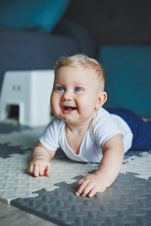 A blue-eyed baby boy in a white bodysuit and blue pants learns to sit. Self-development of the child. The baby is sitting on the floor and smilingの写真素材