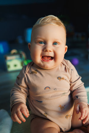 Fair-haired boy with blue eyes baby 11 months in a bodysuit sits on the floor at home and smiles. Children are playing at home.の写真素材