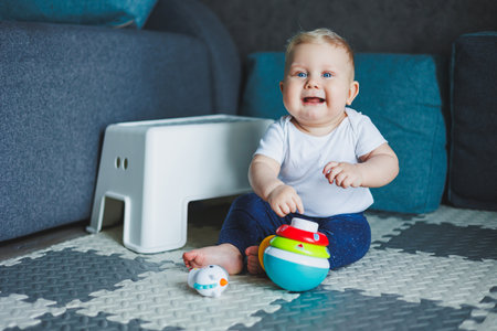 A baby boy in a white bodysuit sits on a rug at home and plays with a plastic pyramid. Educational...