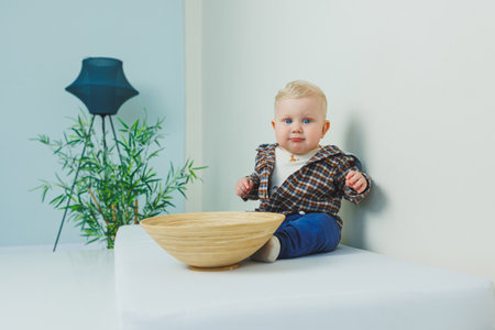 A 9-month-old baby boy in a stylish shirt, pants and sneakers is standing near a support.