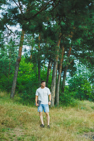 A man in a white T-shirt with a laptop stands in nature and looks into the distance. A man rests in nature from work.の写真素材