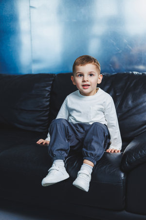 A 3-year-old boy in white raglan and gray pants is sitting on the couch. Stylish boy with a smile on his face. Clothes for boys.の写真素材