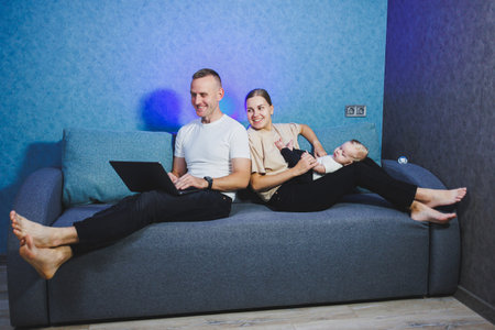 Young family with baby boy playing together at home in living room, parents with baby sitting on sofa, mom and dad holding and kissing baby boy, laughing and having fun.の写真素材